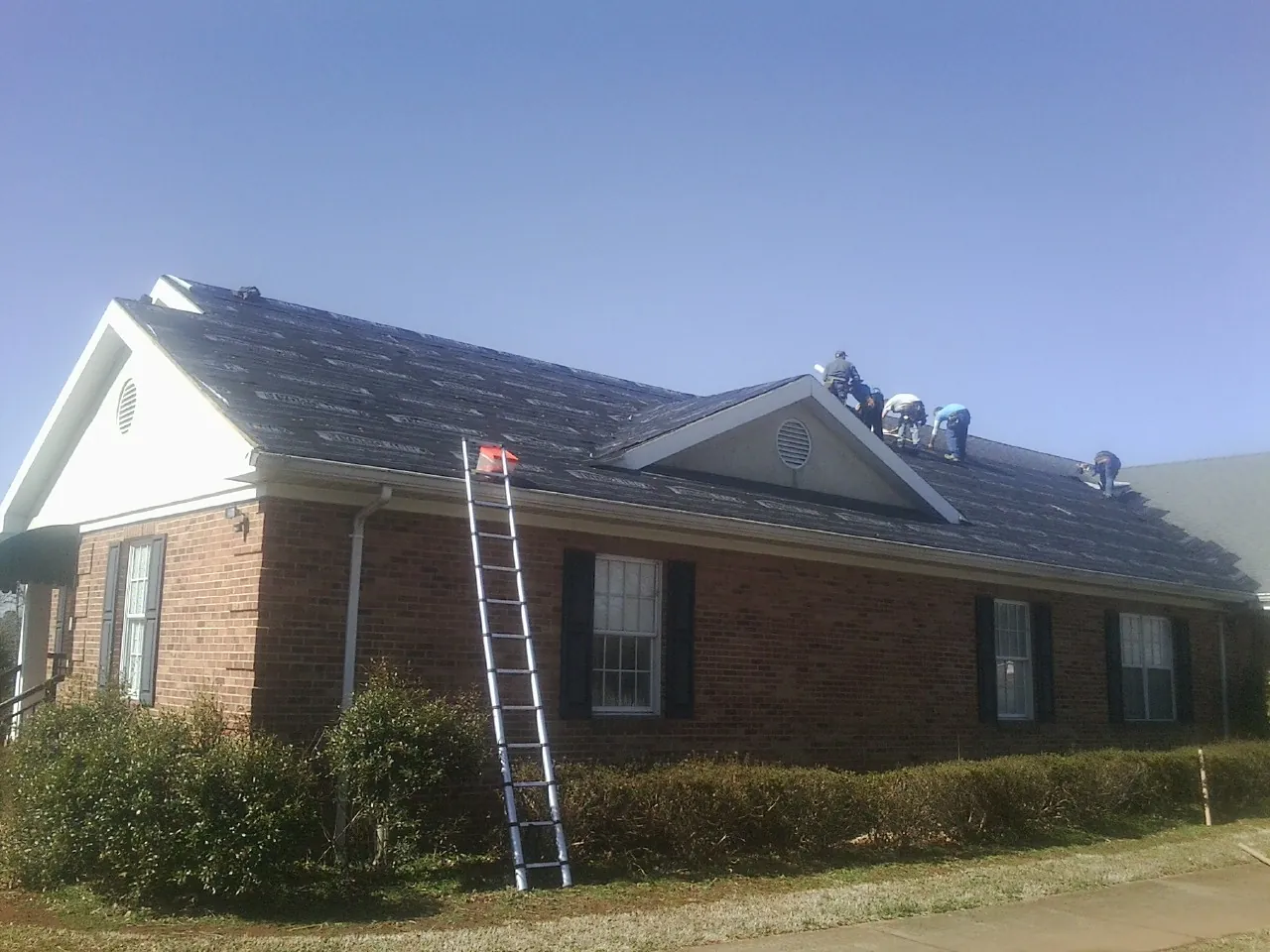 Crew tearing shingles off a brick ranch roof with a ladder set — Emergency Roof Repair work by our crew in Aydlett